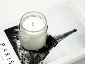 Vela de soja blanca de la marca Velecia, en un frasco de vidrio transparente, colocada sobre un libro de decoración con una imagen de la Torre Eiffel en la portada.