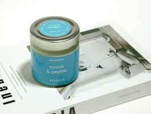 Vela aromática de soja de la marca Velecia, con fragancia de ozono y pepino, en un frasco metálico con tapa plateada y una etiqueta azul claro, colocada sobre un libro de decoración.